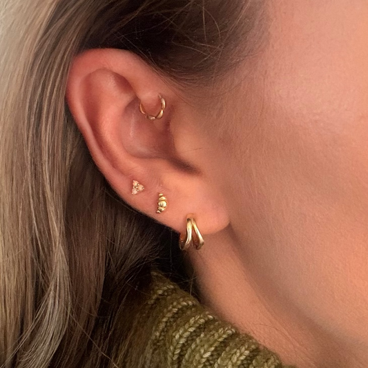 A close up of an ear with a croissant stud in gold, a frilly huggie, a gold hoop and opal gemstone earring