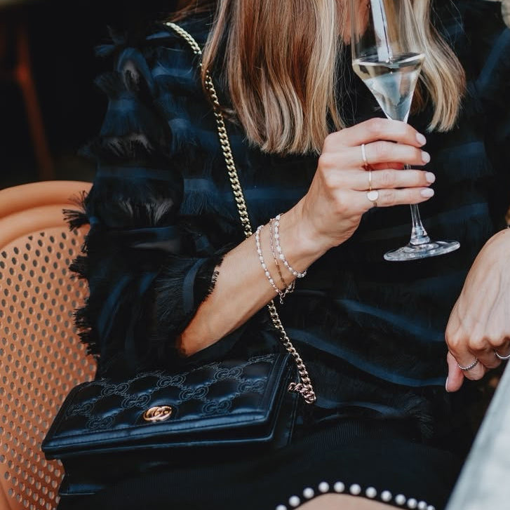 A woman in a black ruffled dress with pearl trim sits at a stone wall cafe, holding a champagne flute and carrying a black quilted shoulder bag with a gold chain strap.