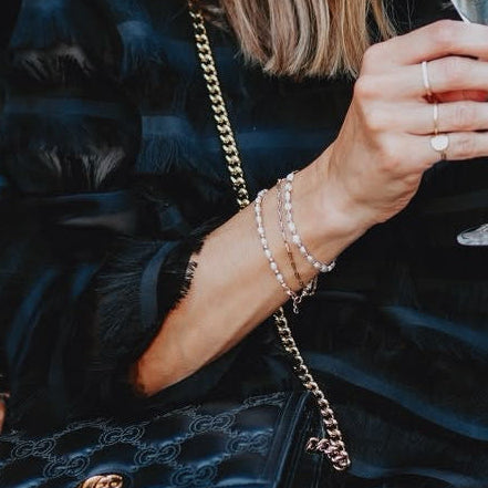A woman in a black ruffled dress with pearl trim sits at a stone wall cafe, holding a champagne flute and carrying a black quilted shoulder bag with a gold chain strap.